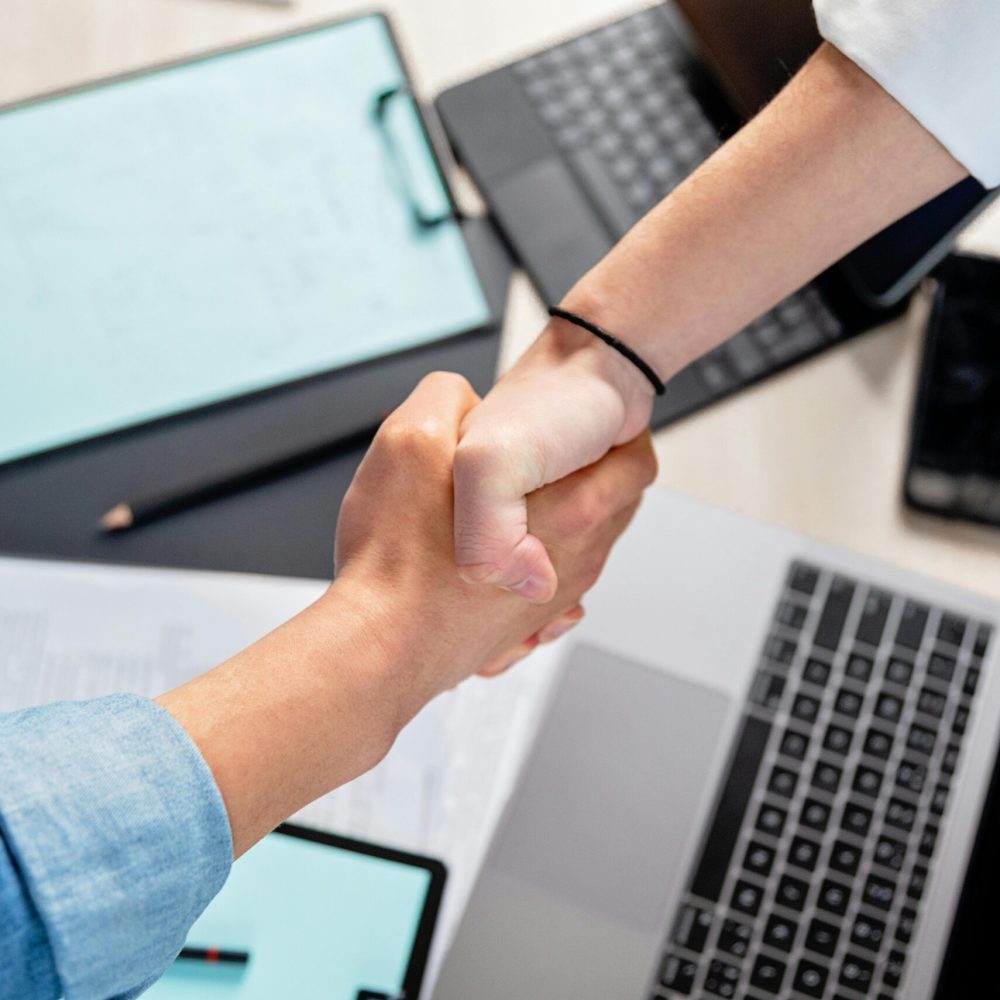 pexels-photo-7989239-7989239 Two people shaking hands over laptops and documents in an office, symbolizing a business deal.